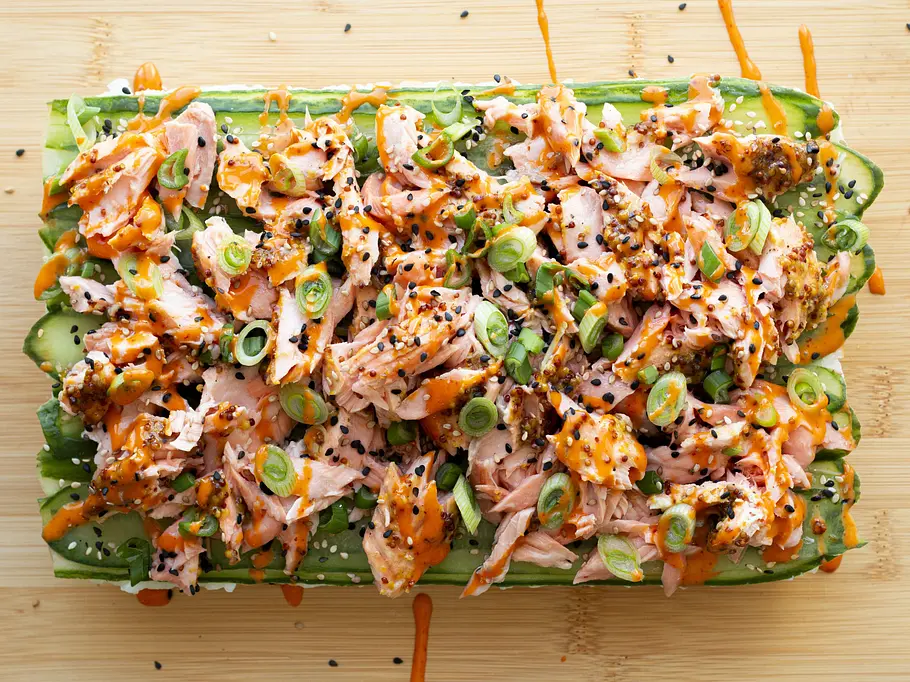 Gestreifter Sushi-Reis mit Gurkenscheiben, darauf zarter Lachs, Sesam, Frühlingszwiebeln und Sriracha-Sauce, angerichtet auf einem Holzbrett.
