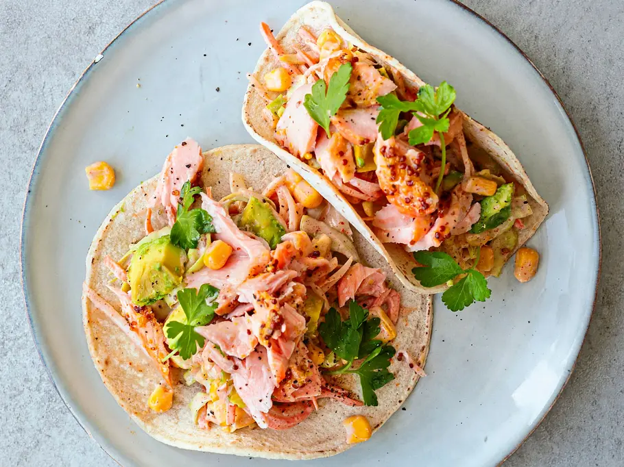 Tacos mit Ofenlachs Zwei Taco-Wraps mit zartem Ofenlachs, Avocado, Mais, frischer Petersilie und Gemüse auf einem hellen Teller.