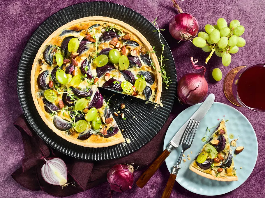 Vegetarischer Zwiebelkuchen mit Trauben Vegetarischer Zwiebelkuchen mit roten Zwiebeln und grünen Trauben auf einem schwarzen Teller, dazu ein Stück auf einem hellblauen Dessertteller mit Besteck.