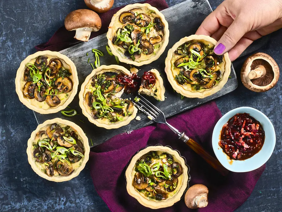 Champignon-Tartelettes mit Speck-Marmelade Kleine Champignon-Tartelettes mit Kräutern auf einer dunklen Platte, dazu ein Schälchen mit Speck-Marmelade und frische Champignons als Dekoration.