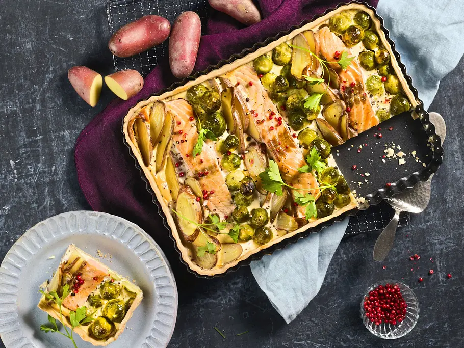 Lachs-Quiche mit Kartoffeln und Rosenkohl Herzhafte Lachs-Quiche mit Kartoffelscheiben und Rosenkohl in einer eckigen Backform auf einem dunklen Untergrund, dazu ein Stück auf einem hellen, geriffelten Teller.