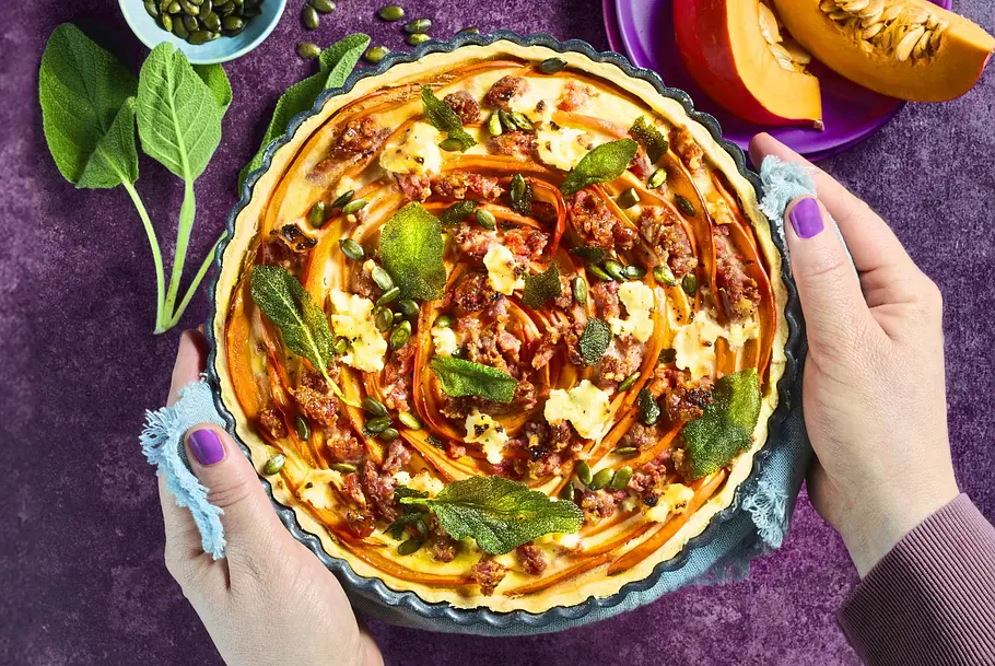 Kürbis-Tarte mit Salsiccia und Salbei-Chips Herzhafte Kürbis-Tarte mit gebratener Salsiccia und knusprigen Salbei-Chips in einer runden Form, daneben Kürbisstücke und Kürbiskerne auf einem kleinen Teller.
