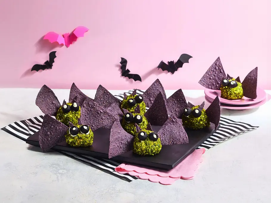 Fledermaus-Bällchen mit Nachos Mehrere grüne, kugelförmige Fledermaus-Bällchen mit auffälligen Augen, dekoriert mit dunklen Dreiecken aus Nachos als Flügel, angerichtet auf einer schwarzen Platte mit gestreifter Serviette.