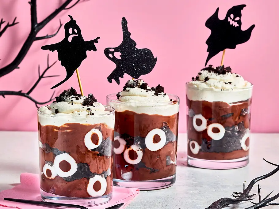 Schaurige Schoko-Schichtcreme Drei Gläser mit schauriger Schoko-Schichtcreme, dekoriert mit weißen Zuckeraugen, Keksstreuseln und Sahne, garniert mit schwarzen Geister-Pieksern, auf rosa Untersetzern angerichtet.