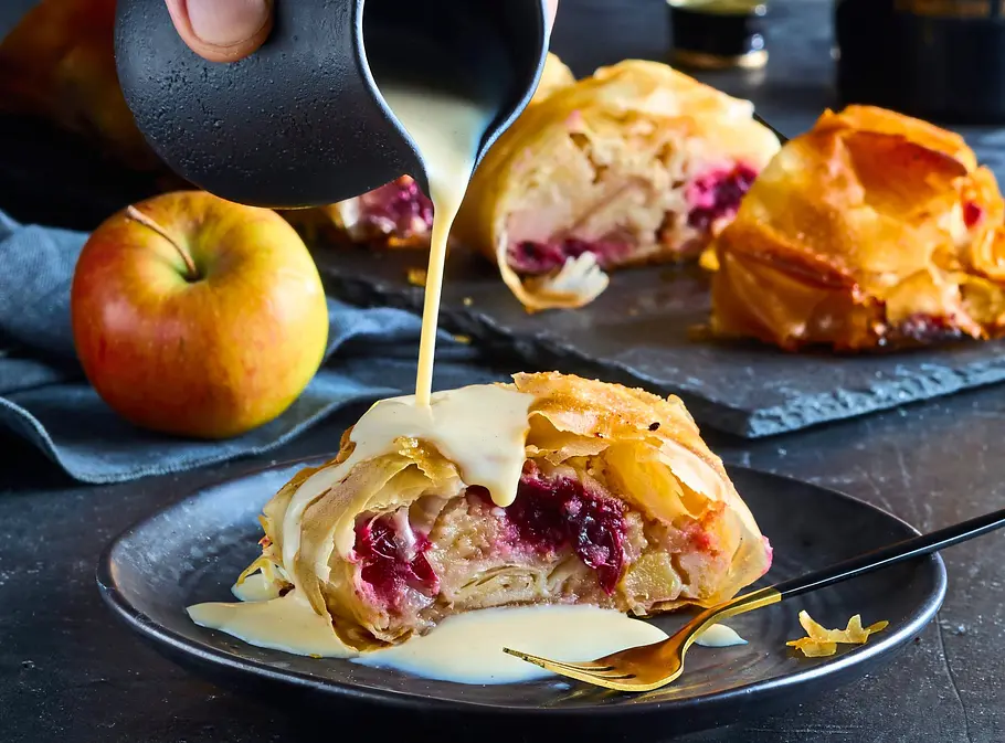 Apfel-Cranberry-Strudel mit Baileys-Vanillesoße Eine Hand gießt helle Sauce über ein Stück Obststrudel. Dahinter stehen ein Apfel, weitere Strudelstücke und eine Baileys-Flasche.