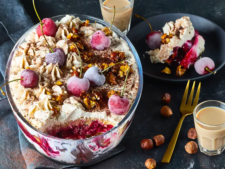 Kirsch-Tiramisu mit Baileys Ein Schichtdessert mit Kirschen und Nüssen steht in einer Glasschale neben einer Portion auf einem Teller und zwei Gläsern.