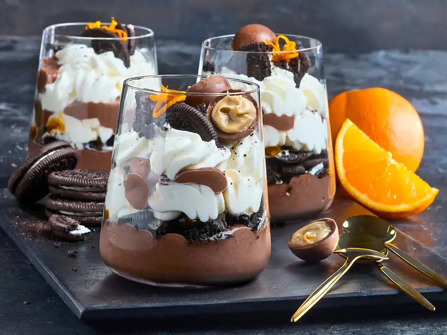 Mocca-Trifle „Baileys & Oreo“ Drei Gläser mit Schokodessert, Sahne und Keksen stehen neben einer Orange, Pralinen und goldfarbenen Löffeln.