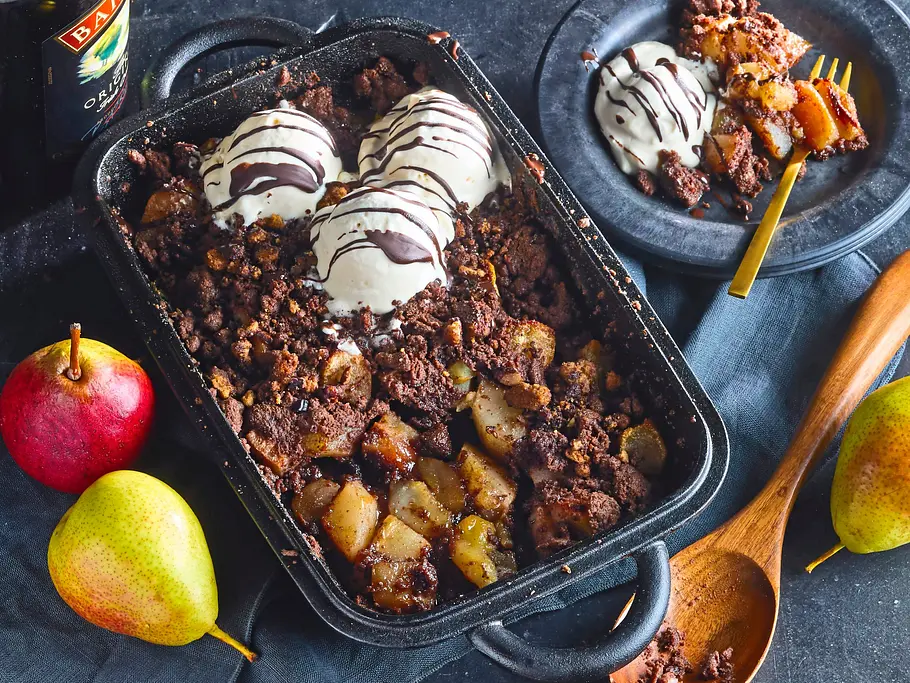 Baileys-Eis auf Birnen-Crumble Birnencrumble mit Eis und Schokosoße steht in einer Backform neben einem Dessertteller, Birnen, Holzlöffel und einer Baileys-Flasche.