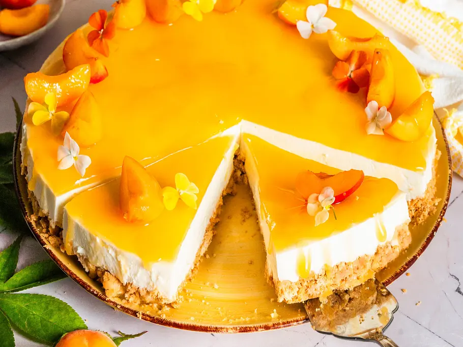 Buttermilch-Zitronen-Kuchen mit Aprikosenspiegel Runder Buttermilch-Zitronen-Kuchen mit Aprikosenspiegel, verziert mit Aprikosenstücken und essbaren Blüten auf einem goldenen Teller, ein Stück angeschnitten, daneben frische Aprikosen und eine gelbe Schüssel.