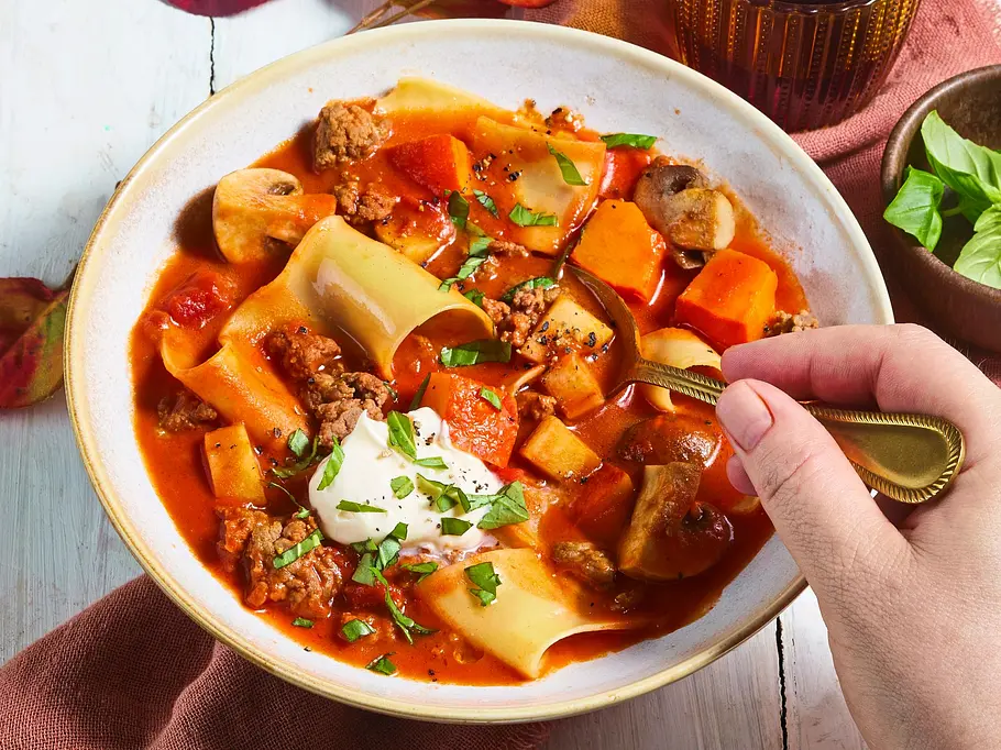 Herbstliche Lasagne-Suppe mit Champignons und Kürbis Eine Hand hält einen Löffel in einer Schale mit Nudeln, Hackfleisch, Pilzen und roter Soße. Daneben liegen Herbstblätter.
