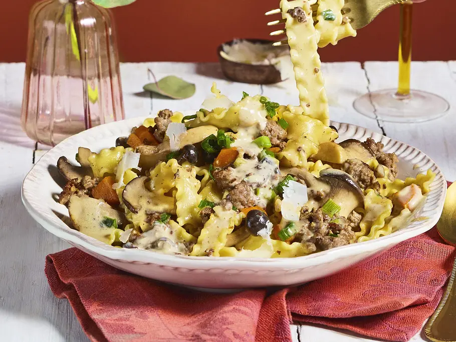 Pasta mit Jäger-Bolo Eine Hand hebt Nudeln mit einer Gabel vom Teller mit Pilzen, Hackfleisch und Käse. Dahinter stehen eine Vase und ein Weinglas.