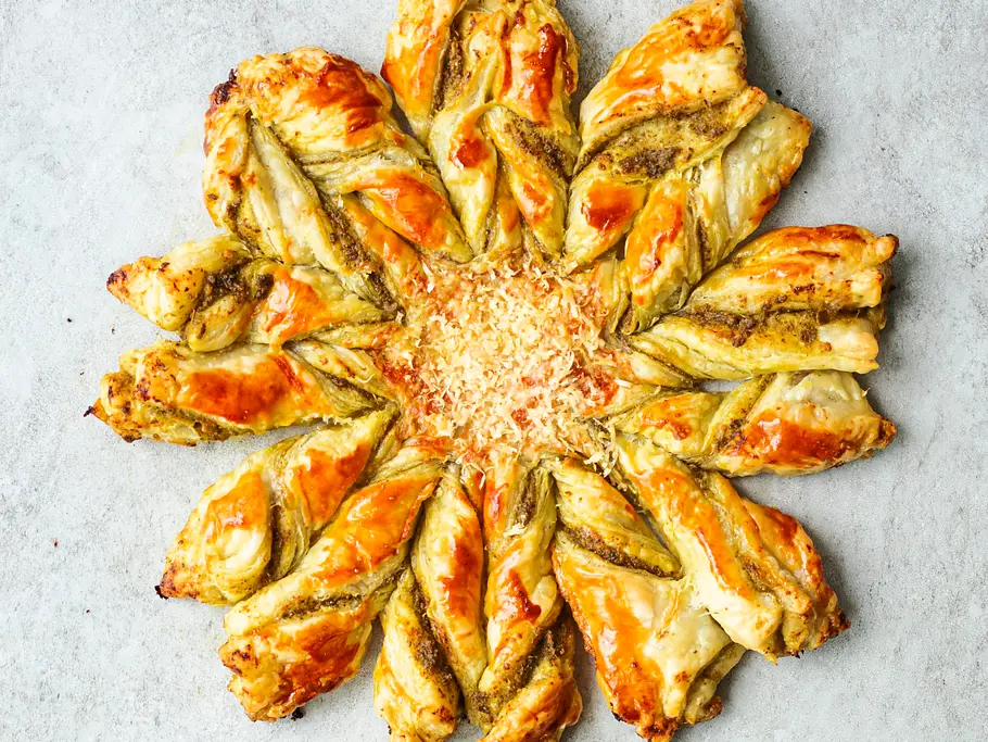 Goldbraune Blätterteigblume mit gedrehten, knusprigen „Blütenblättern“ und geriebenem Käse in der Mitte, serviert auf heller Unterlage.