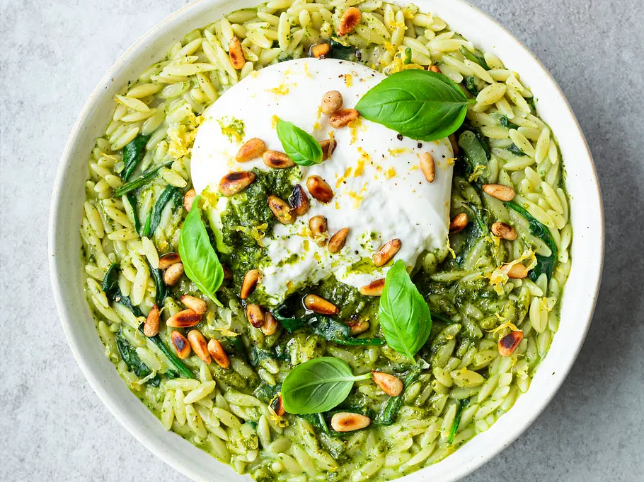 Green Orzotto Cremiges grünes Orzotto aus Risoni-Nudeln mit Spinat und Pesto, garniert mit Burrata, gerösteten Pinienkernen und frischem Basilikum in einer weißen Schale.