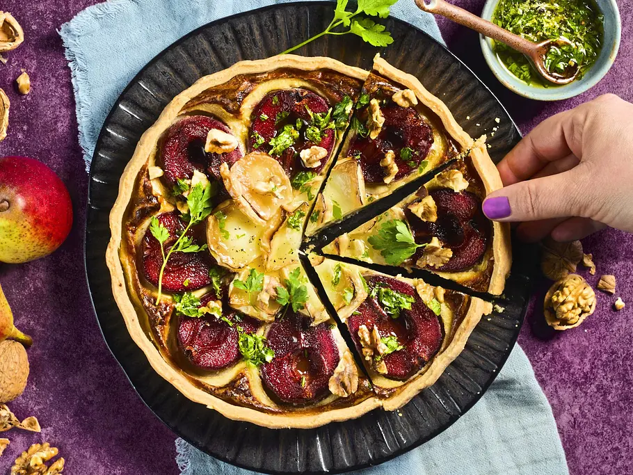 Tarte mit Rotwein-Birnen und Ziegenkäse Tarte mit Rotwein-Birnen und Ziegenkäse auf einem schwarzen Teller, garniert mit frischen Kräutern und Walnüssen, serviert auf einem hellblauen Stofftuch.
