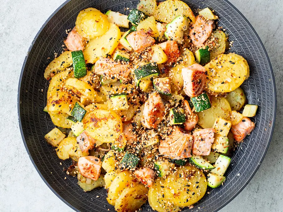 Bratkartoffeln mit Lachs und Zucchini Bratkartoffeln mit zarten Lachsstücken und gebratener Zucchini, bestreut mit Sesam, serviert in einer dunklen Pfanne.
