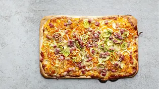 Knuspriger Flammkuchen auf eckigem Teigboden, belegt mit Speckwürfeln, Lauchringen und cremiger Carbonara-Soße. - Foto: Simone Neufing/MOE‘S QUICK & EASY FOOD