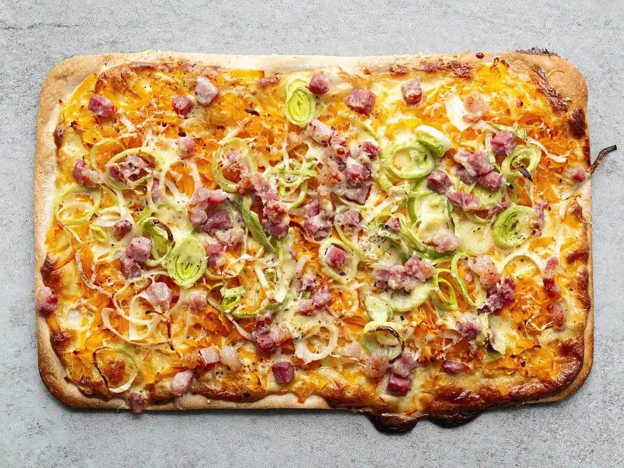 Carbonara-Flammkuchen Knuspriger Flammkuchen auf eckigem Teigboden, belegt mit Speckwürfeln, Lauchringen und cremiger Carbonara-Soße.