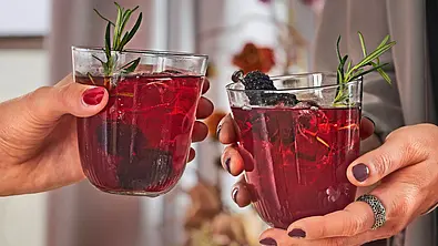 Zwei Gläser mit einem tiefroten Cocktail, garniert mit Brombeeren und einem Zweig frischem Rosmarin. Die Gläser werden von zwei Personen in elegantem Ambiente gehalten, im Hintergrund brennen Kerzen. - Foto: LECKER @ Bauer Media Group