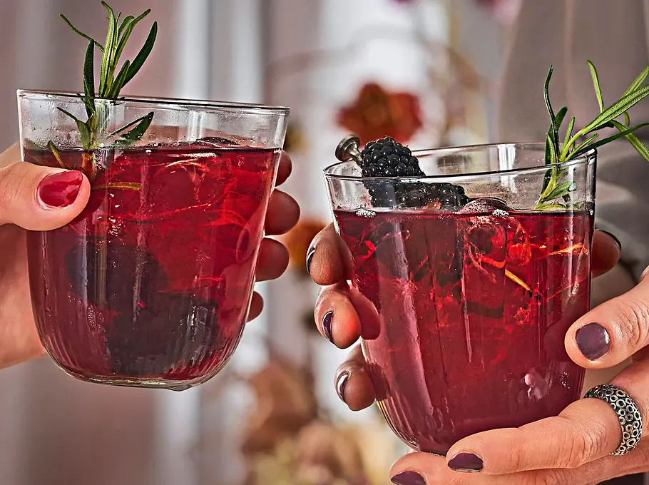 Zwei Gläser mit einem tiefroten Cocktail, garniert mit Brombeeren und einem Zweig frischem Rosmarin. Die Gläser werden von zwei Personen in elegantem Ambiente gehalten, im Hintergrund brennen Kerzen.