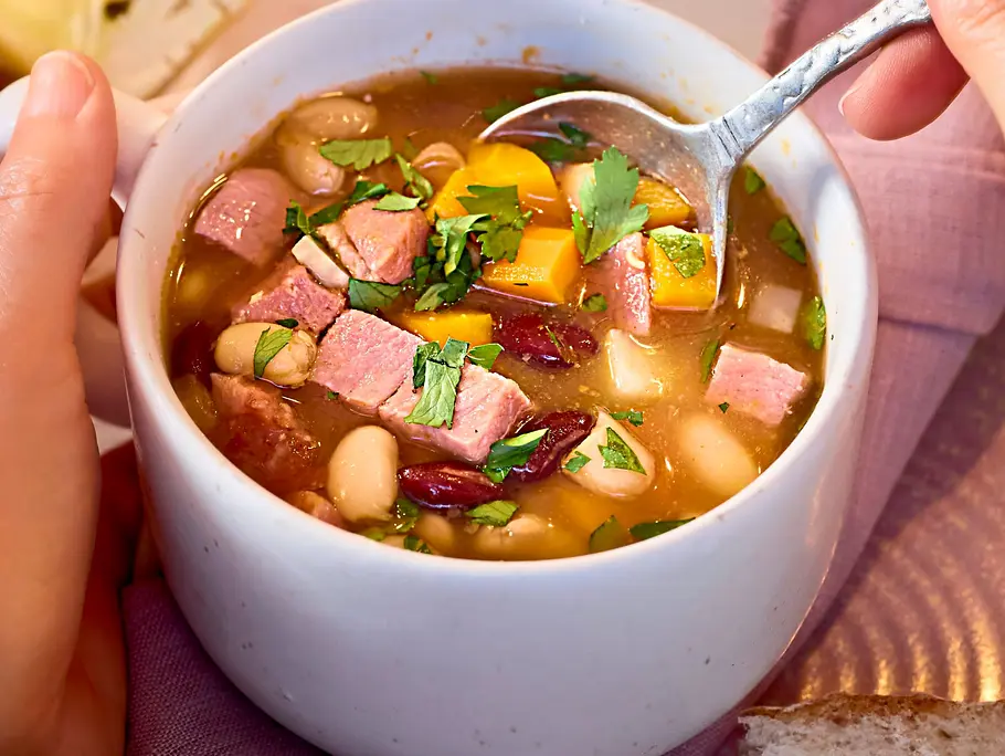 Bunte Bohnensuppe mit Kasselerwürfeln, serviert in einer weißen Tasse auf einem pastellfarbenen Teller mit einem Stück Brot daneben.