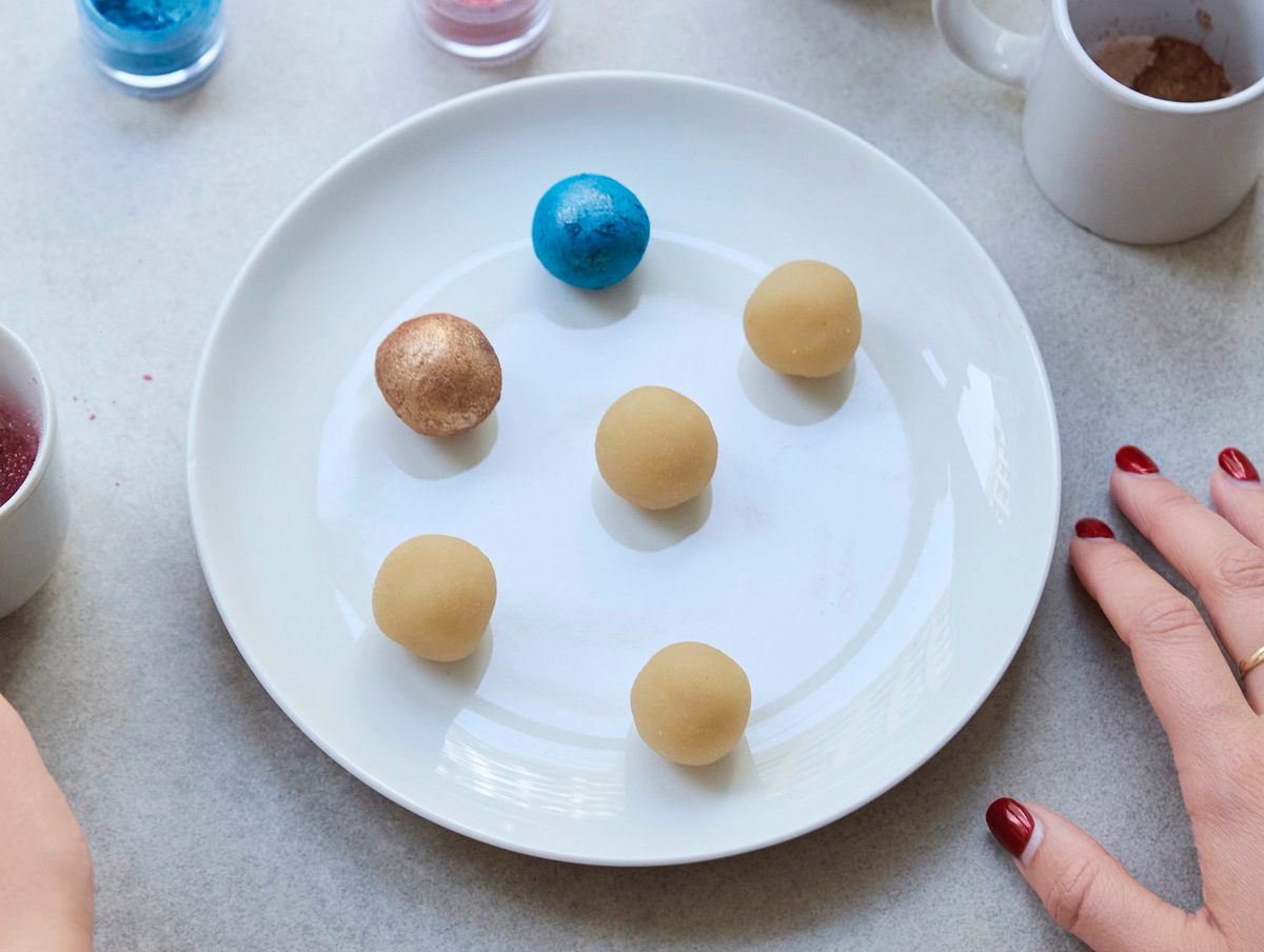  Verschiedene Marzipankugeln auf einem Teller, einige bereits farbig bemalt in Blau, Gold und Rosa, daneben kleine Schälchen mit essbarem Puder und Pinsel zum Verzieren.