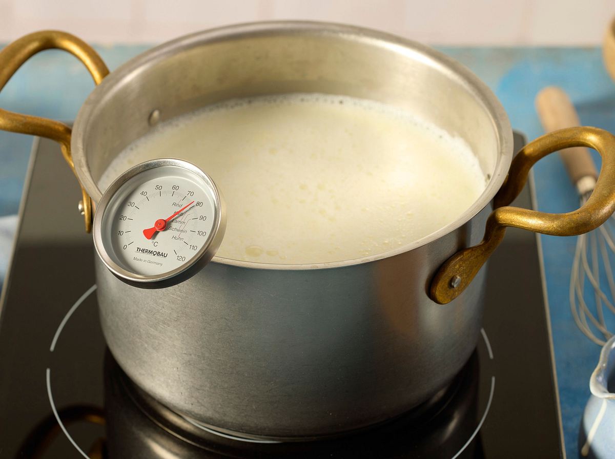 Milch und Sahne in einem Topf auf dem Herd erhitzt, ein Küchenthermometer zeigt 80 °C, die Mischung ist kurz vorm Kochen.