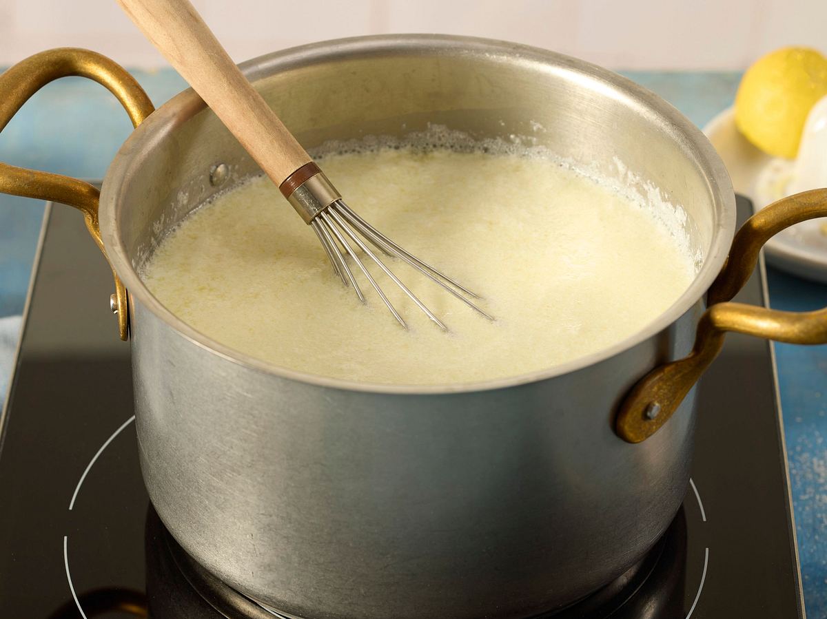 Die Milch-Sahne-Mischung im Topf zeigt eine geronnene, ausgeflockte Konsistenz, verursacht durch die Zugabe von Zitronensaft; ein Schneebesen liegt im Topf.