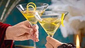 Elegant servierte Vesper-Martinis in klassischen Cocktailgläsern, dekoriert mit einer Zitronenzeste, die angestoßen werden; auf einem festlich gedeckten Tisch mit goldfarbenem Tablett und grünen Kerzen. - Foto: LECKER @ Bauer Media Group