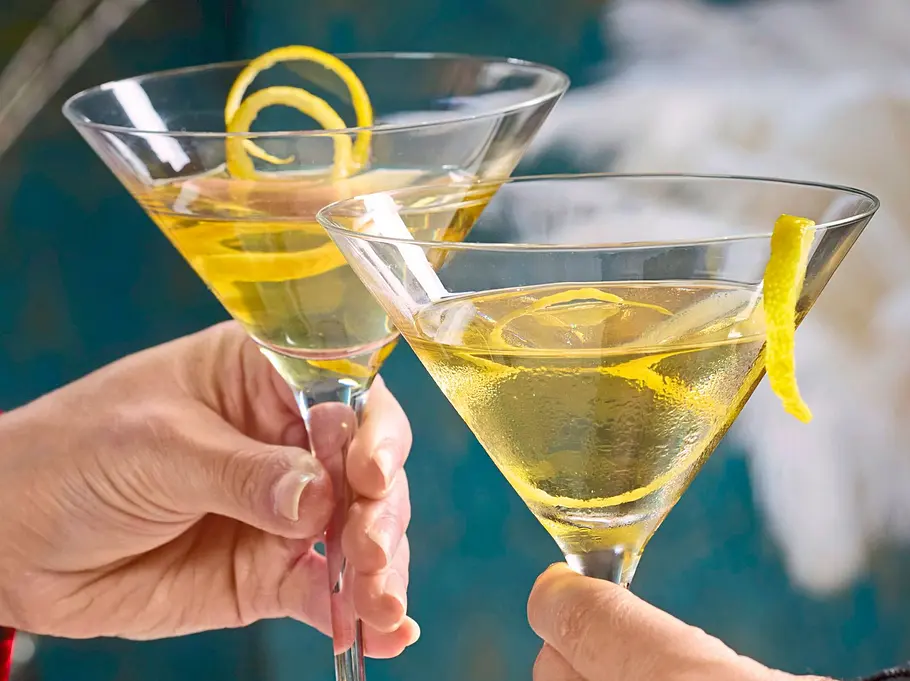 Elegant servierte Vesper-Martinis in klassischen Cocktailgläsern, dekoriert mit einer Zitronenzeste, die angestoßen werden; auf einem festlich gedeckten Tisch mit goldfarbenem Tablett und grünen Kerzen.