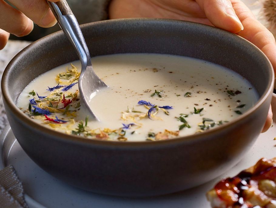 Cremige Bergkäsesuppe in einer dunklen Schale, verfeinert mit bunten Blüten und frischen Kräutern, serviert auf einem hellen Teller.