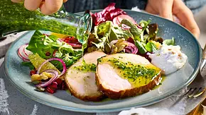 Zwei Scheiben saftiger Kasselerlachs auf einem hellen Teller, serviert mit bunter Blattsalatmischung, roten Zwiebeln, Nüssen und frischer Kren-Creme, dazu grüne Kräutersauce. - Foto: LECKER @ Bauer Media Group