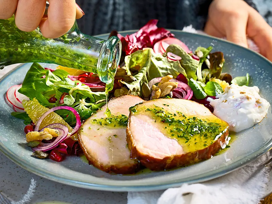 Zwei Scheiben saftiger Kasselerlachs auf einem hellen Teller, serviert mit bunter Blattsalatmischung, roten Zwiebeln, Nüssen und frischer Kren-Creme, dazu grüne Kräutersauce.
