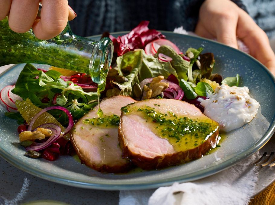 Zartes Honig-Kasseler in Scheiben, mit Kräuterpesto beträufelt, serviert auf einem grünen Teller mit frischem Blattsalat und cremigem Meerrettich-Dip.