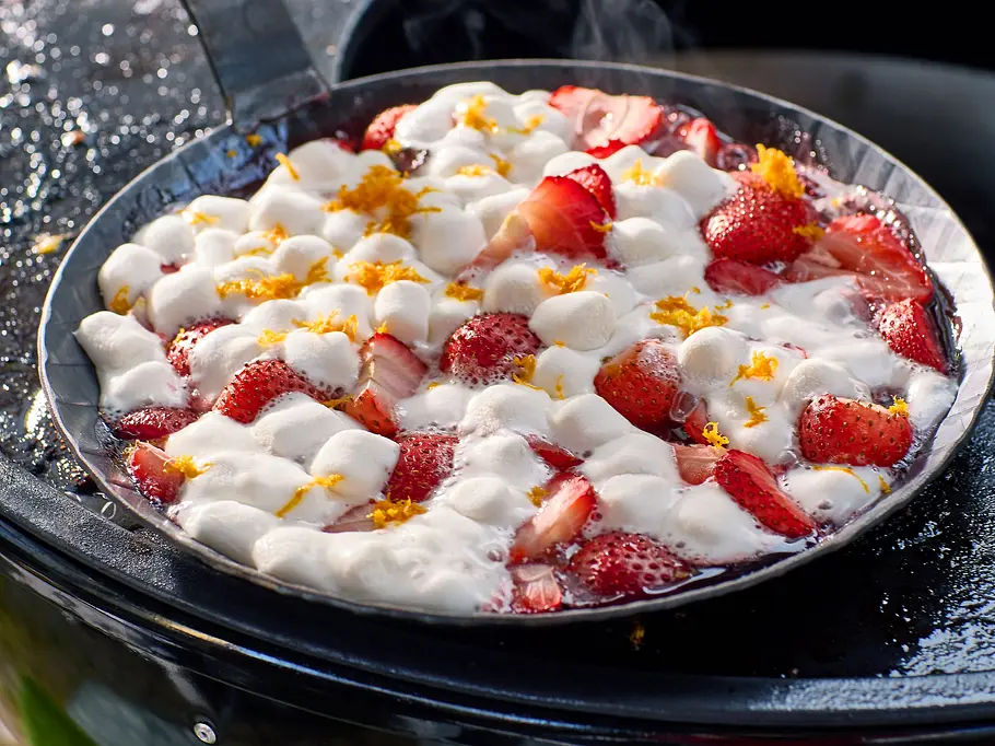 Marshmallow-Erdbeeren vom Grill Auf einer Plancha-Platte steht eine Grillpfanne mit geschmolzenen Marshmallow, kleingeschnittenen Erdbeeren und Orangenabrieb