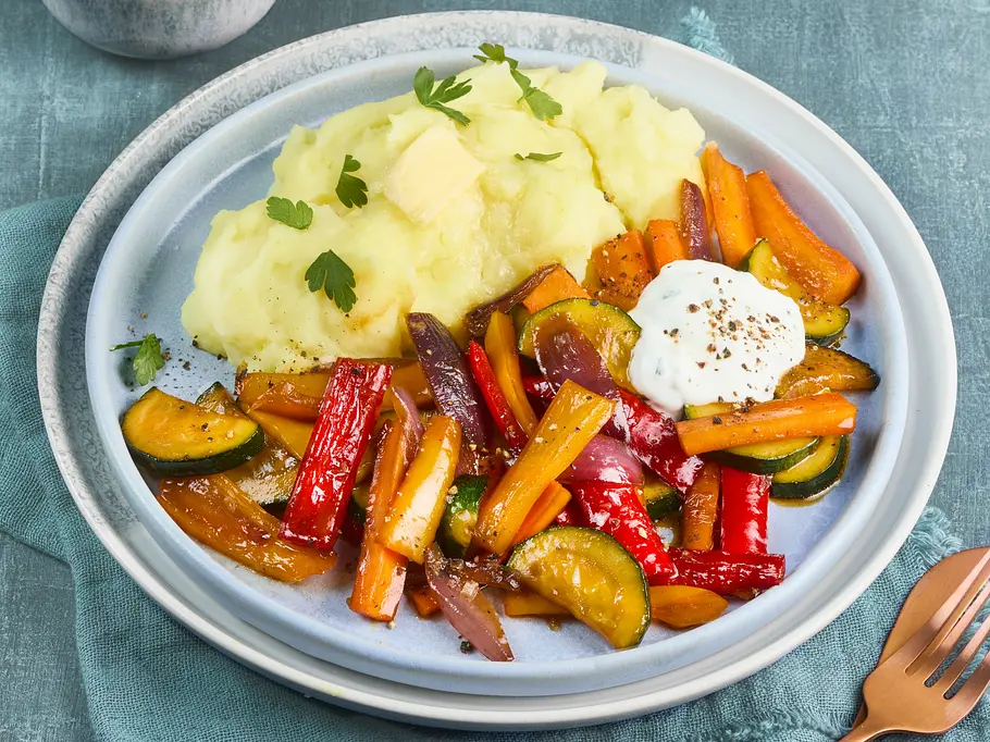 Honig-Gemüse mit Kartoffelpüree Honig glasiertes buntes Ofengemüse mit Paprika, Zucchini und Karotten, serviert auf einem hellen Teller mit cremigem Kartoffelpüree und Kräuterdip, angerichtet auf einer hellblauen Stoffserviette.