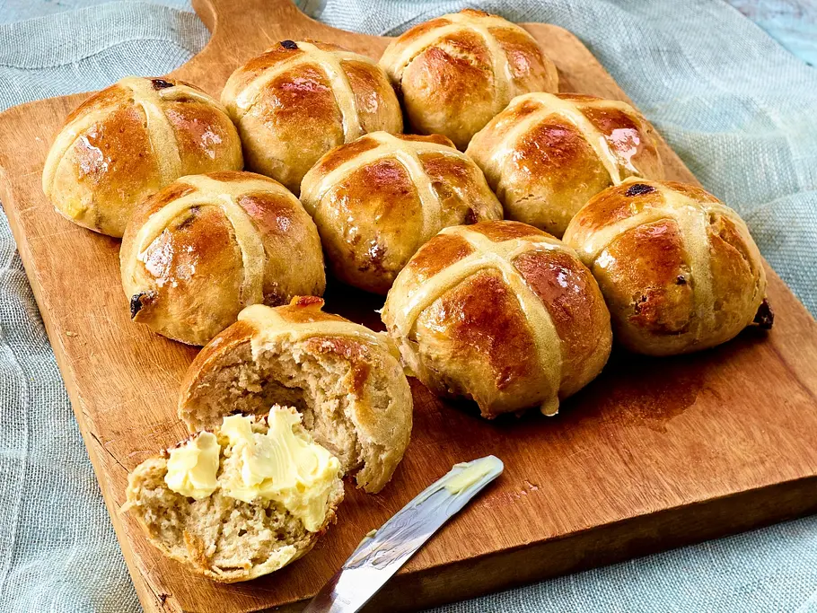 Ein Schneidebrett mit fertig gebackenen Hot Cross Buns. Eines ist aufgeschnitten und mit Butter bestrichen. Das Messer liegt daneben. 