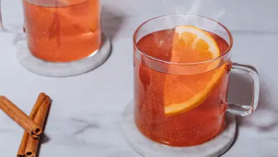 Auf einem hellen Untergrund stehen zwei Tassen aus Glas, gefüllt mit Hot Glitter Spritz, man sieht etwas Dampf aufsteigen, die Flüssigkeit ist leuchtend rot-orange und glitzert - Foto: Craft Circus GmbH, KI-optimiert