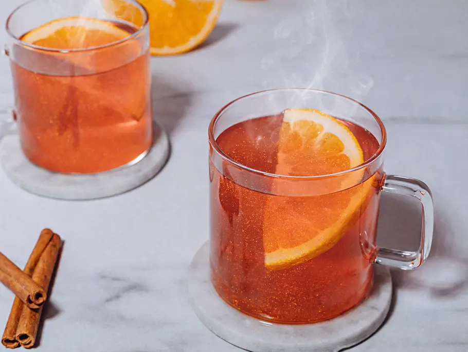 Auf einem hellen Untergrund stehen zwei Tassen aus Glas, gefüllt mit Hot Glitter Spritz, man sieht etwas Dampf aufsteigen, die Flüssigkeit ist leuchtend rot-orange und glitzert