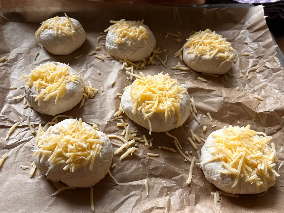 7 Teiglinge liegen mit Käse bestreut auf einem mit Backpapier ausgelegtem Backblech