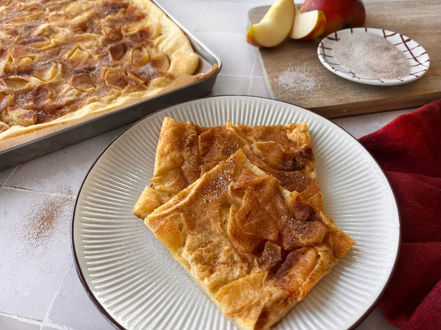 Apfelpfannkuchen vom Blech Apfelpfannkuchen vom Blech mit Zimt und Zucker, angerichtet auf einem Teller mit frischen Äpfeln.