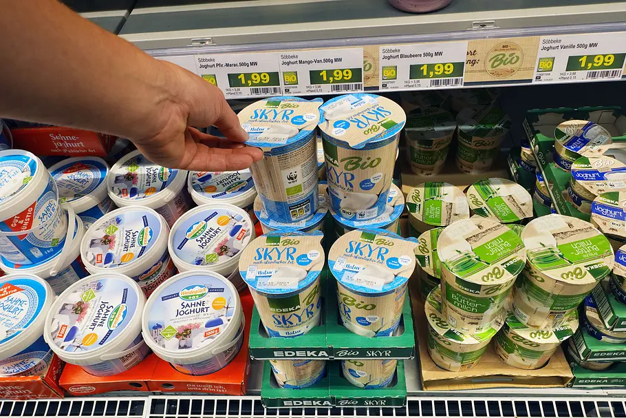Hand greift im Supermarktregal nach einem Becher Skyr zwischen anderen Joghurtprodukten; Preisschilder und verschiedene Sorten sind sichtbar.
