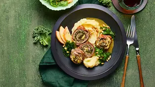 Drei gerollte Involtini mit Grünkohl und Caprese-Füllung auf cremigem Kartoffelpüree, angerichtet auf einem dunklen Teller, dazu Apfelscheiben und frischer Grünkohl, auf grünem Untergrund mit Besteck und grünem Stoffserviette daneben. - Foto: LECKER @ Bauer Media Group