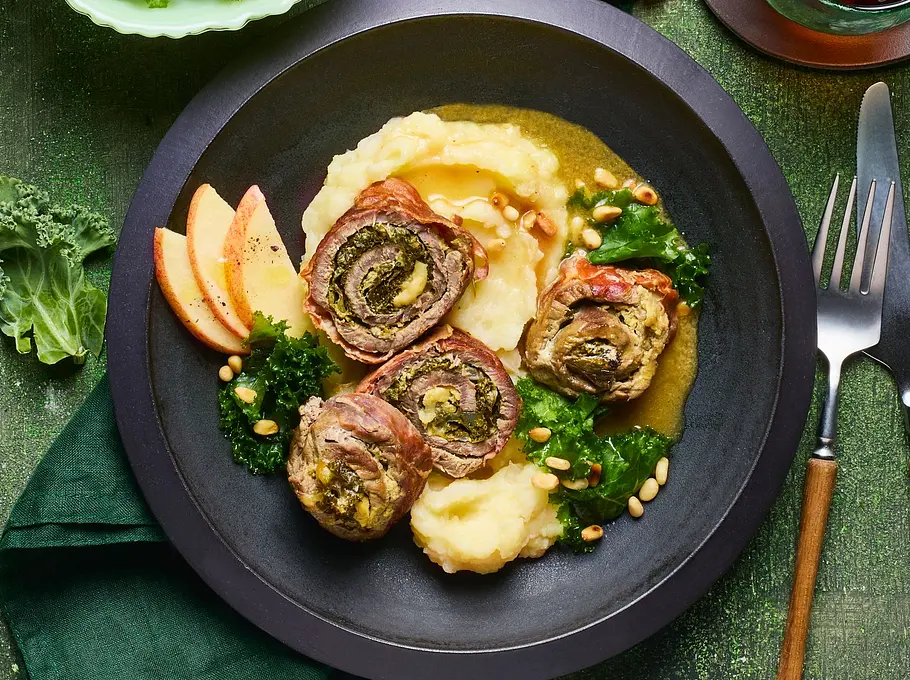 Drei gerollte Involtini mit Grünkohl und Caprese-Füllung auf cremigem Kartoffelpüree, angerichtet auf einem dunklen Teller, dazu Apfelscheiben und frischer Grünkohl, auf grünem Untergrund mit Besteck und grünem Stoffserviette daneben.