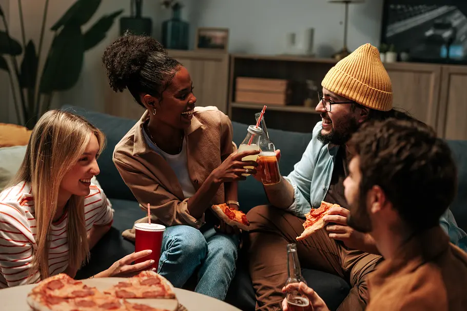 Eine vierköpfige Freundesgruppe sitzt bei warmen Licht zusammen, alle stoßen mit diversen Kaltgetränken an und teilen sich eine Pizza