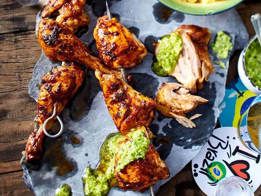 Saftige, kräftig gewürzte Jerk-Chicken-Stücke mit Grillstreifen, angerichtet auf einer Schieferplatte und serviert mit einer frischen grünen Soße.