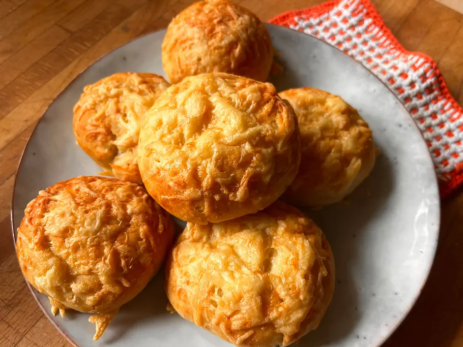 6 goldbraune Käsebrötchen liegen auf einem Teller mit orangenem Küchentuch. 