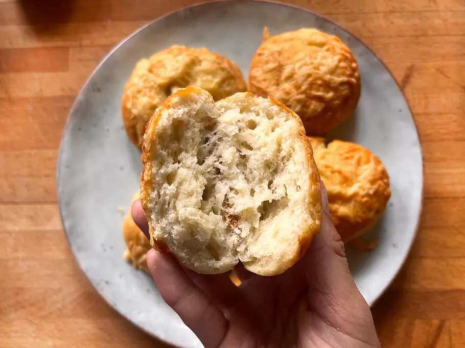 Ein aufgebrochenes Käsebrötchen in der Hand, im Hintergrund ein Teller mit weiteren Käsebrötchen