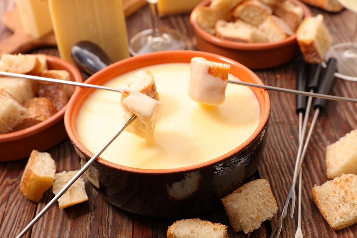 Käsefondue-Topf mit flüssigem Käse auf einem gedeckten Tisch. Darüber drei Fondue-Gabeln mit Brot 