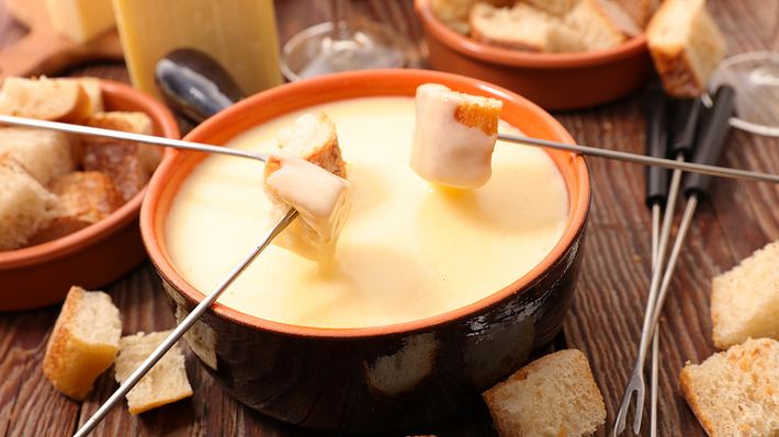 Käsefondue-Topf mit flüssigem Käse auf einem gedeckten Tisch. Darüber drei Fondue-Gabeln mit Brot  - Foto: iStock/margouillatphotos