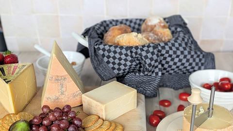 Verschiedene Käsesorten, Trauben, Feigen und Cracker liegen auf einem Holzbrett, dahinter Gebäck in einem Brotkorb. - Foto: Emily Beckmann / LECKER.de